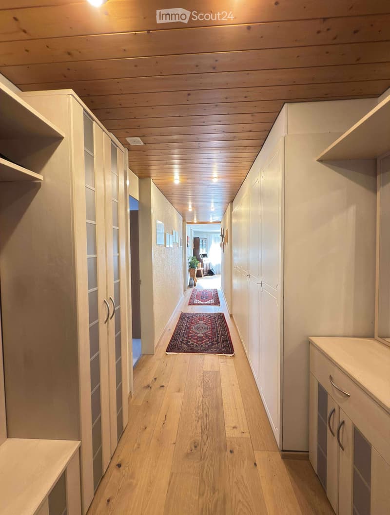 A long, well-lit hallway with wooden flooring, white built-in cabinets, and two patterned rugs placed on the floor. The ceiling features exposed wooden beams and recessed lighting.