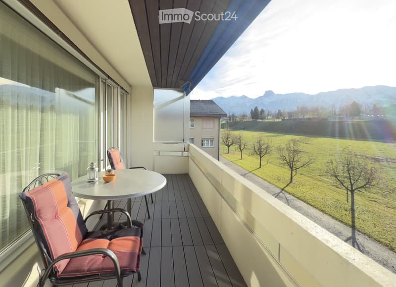 Balcony with a round table, two chairs, and a scenic view of a green field and mountains.