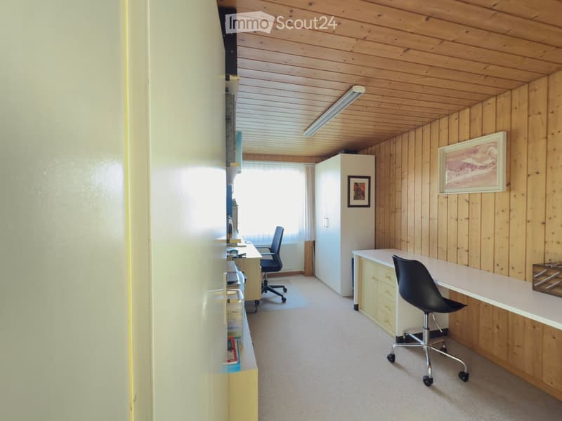 Bright, wooden-paneled room with two desks, chairs, a cabinet, and framed pictures on the wall.
