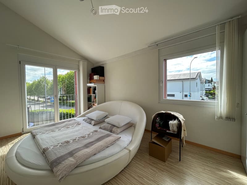 Modern bedroom with a round bed, nightstand, shelf, and large windows with curtains. The room features light-colored walls and a view of the street.