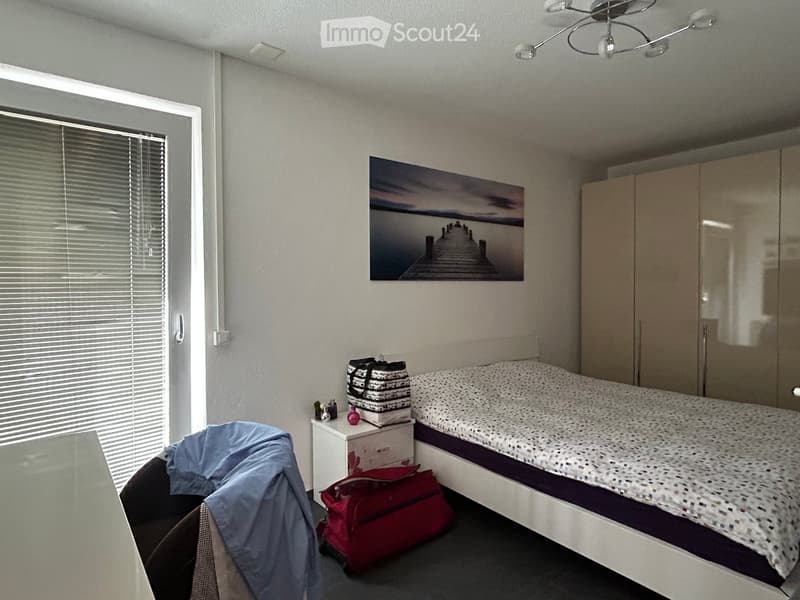 A modern bedroom with a bed, nightstand, chair, and wardrobe. A large picture frame hangs on the wall, and a suitcase is placed on the floor.