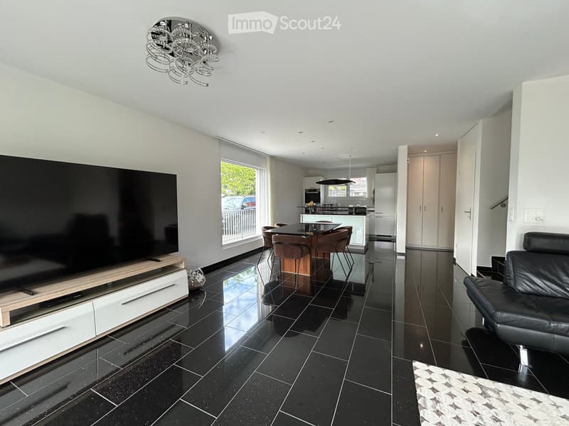 Modern living room with black tiled flooring, large flat-screen TV on a wooden stand, leather sofa, coffee table, dining area with chairs and island, and a view of the kitchen area.
