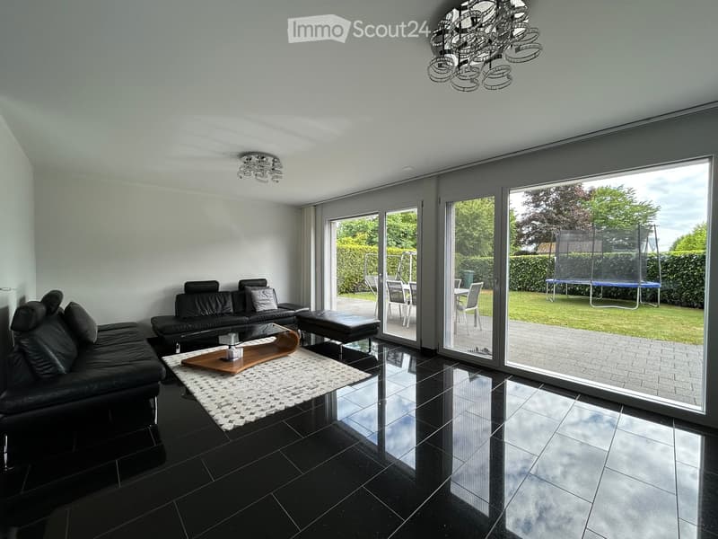 Modern living room with black tiled flooring, large sliding glass doors leading to a patio, a black leather sofa, a wooden coffee table, and a small ottoman. The room features a chandelier and a view of a garden with a trampoline.