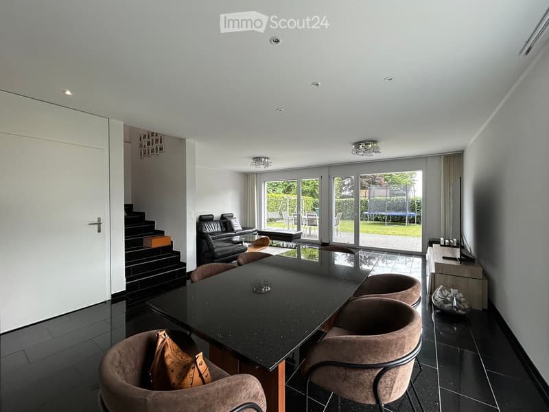 Modern living and dining area with a sleek black dining table, four brown chairs, a black leather sofa, a coffee table, and a TV stand with a flat-screen TV. The space features large glass doors leading to a garden, recessed ceiling lighting, and a stairc