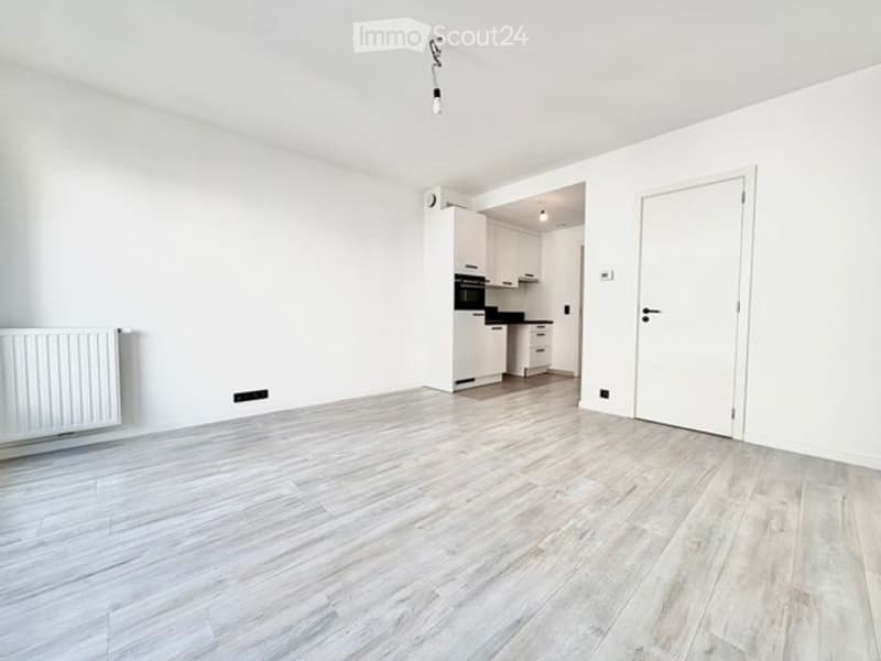 Empty room with white walls, light wooden flooring, and a radiator on one wall. A doorway leads to a small kitchen area with white cabinets and a black oven.