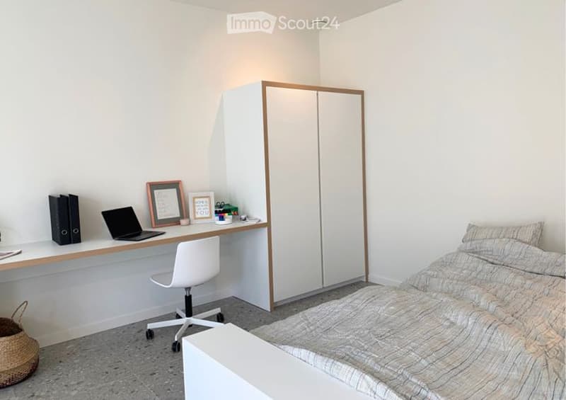 A modern, well-lit bedroom with a desk and a bed. The desk features a laptop, books, and a framed picture, while the bed has a patterned duvet and pillows. A white wardrobe with wooden trim is positioned against the wall.