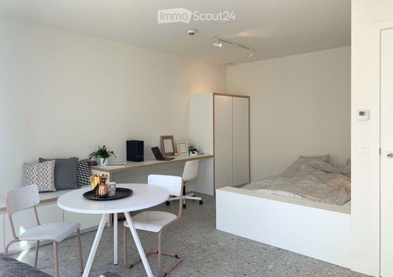 A modern, minimalist bedroom with a bed, desk, and small round table. The room features white walls, a grey floor, and contemporary furniture including a white desk chair, a small round table with chairs, and a potted plant on the desk.