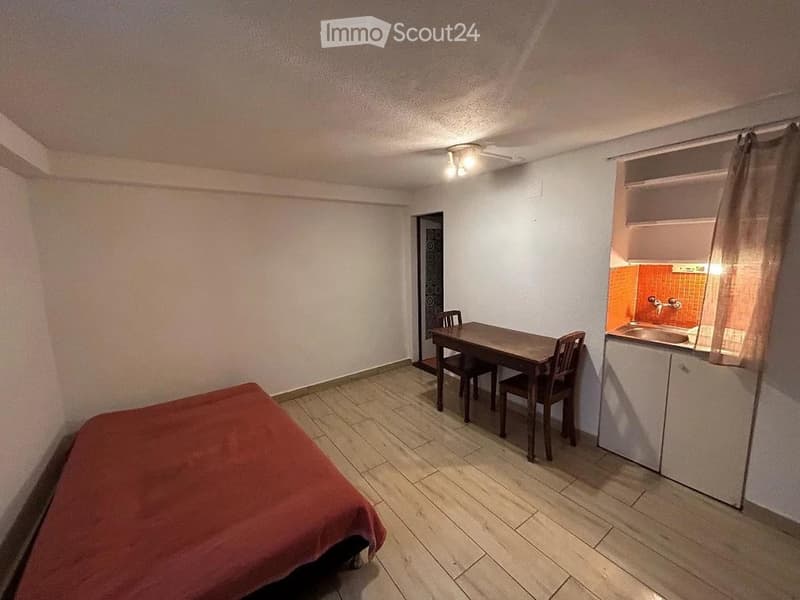 A small, furnished room with a bed, a dining table with chairs, a sink, and a cabinet.
