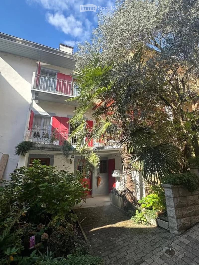 Two-story white building with red shutters, balconies, and lush greenery including palm trees and shrubs.