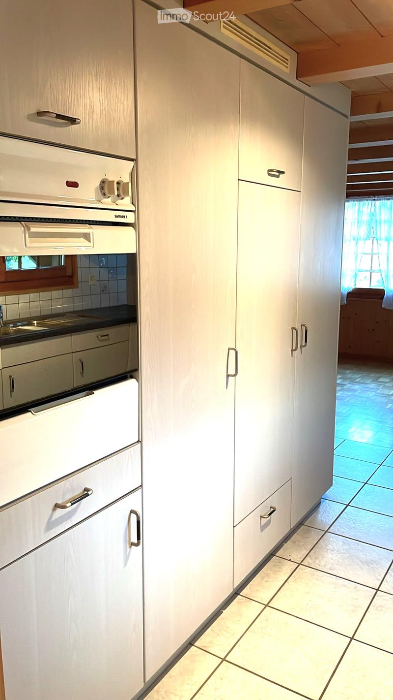 Modern kitchen with white cabinets, stainless steel oven, and tiled floor. A glimpse of a living area with a sofa is visible through an open doorway.