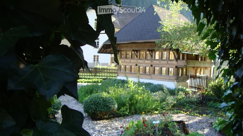 Garden with gravel path, shrubs, and flowers, with a wooden fence and traditional house in the background.