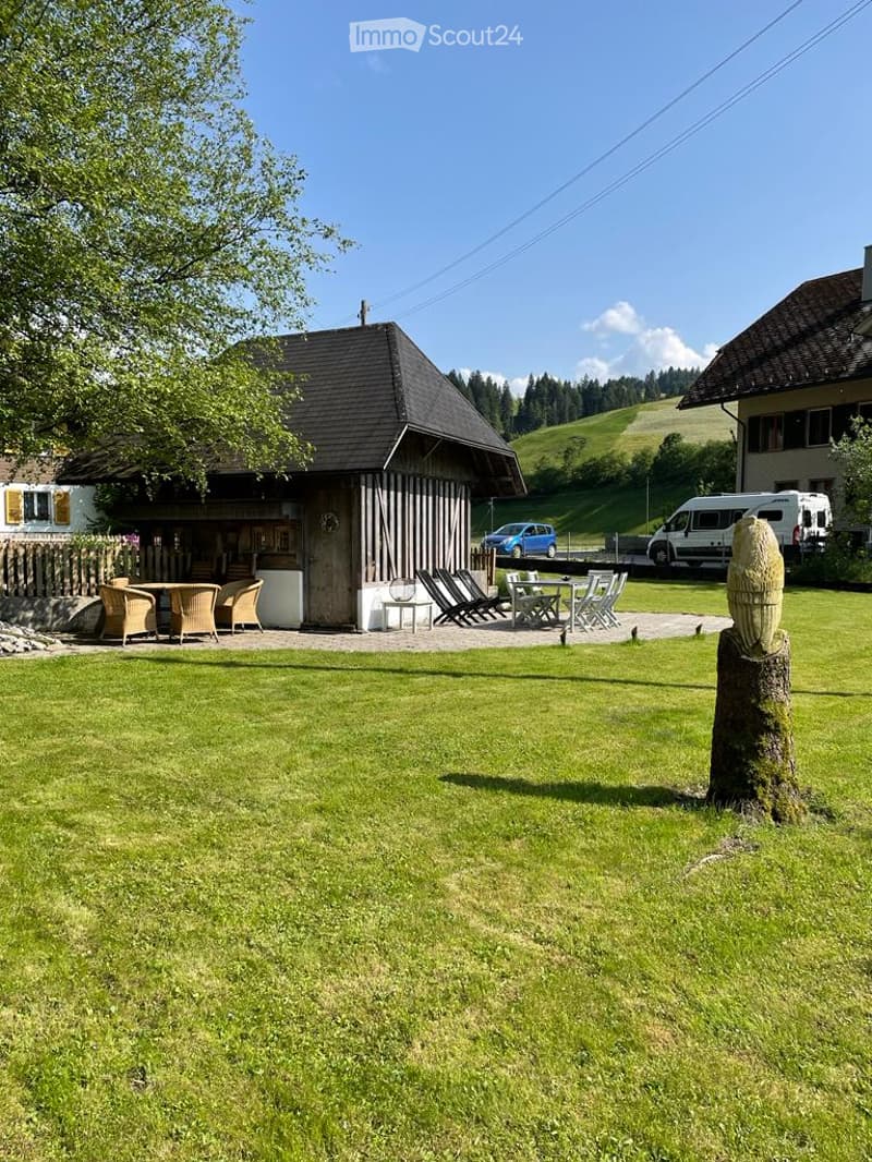 Lush green lawn with outdoor seating area, including chairs and tables, near a wooden structure and parked vehicles.