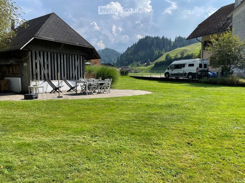 Lush green lawn with a patio area featuring a dining table, chairs, and lounge chairs. A wooden barn and a parked van are visible in the background, with a scenic mountain view.