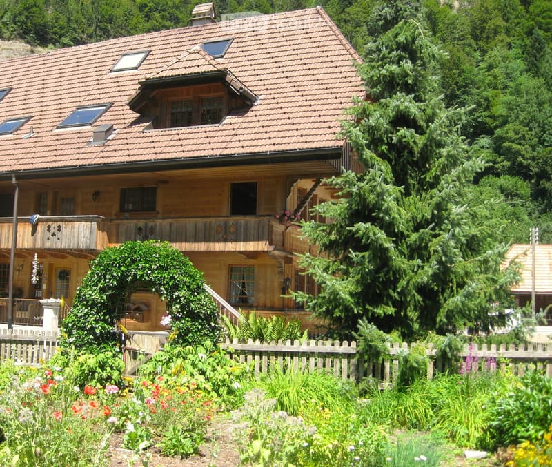 Two-story wooden house with a tiled roof, multiple windows, and a balcony, surrounded by a garden with colorful flowers and a wooden fence.