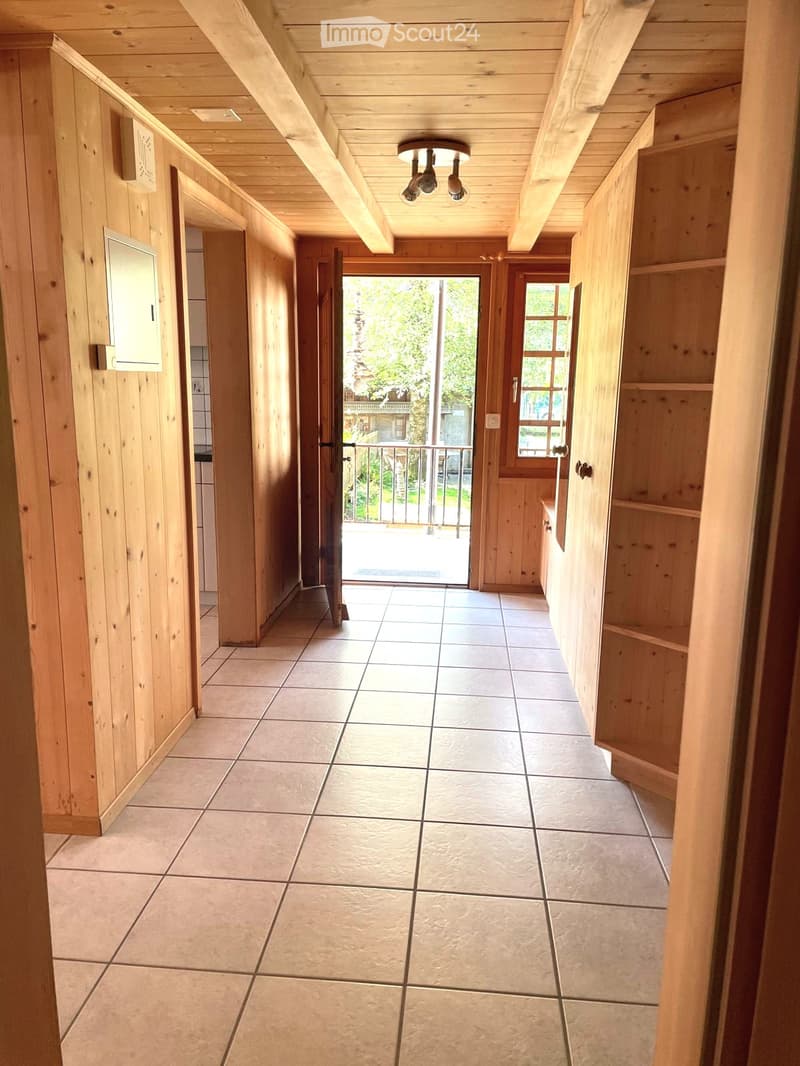 Wooden-paneled hallway with tiled floor, exposed wooden beams, and recessed lighting. Features built-in wooden shelving and open doors leading to other rooms and an outdoor area.
