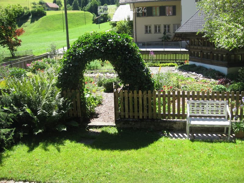 Well-maintained garden with a white bench, wooden fence, and lush greenery, surrounded by a rural landscape with houses and hills in the background.