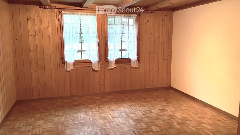 Empty room with wooden walls and herringbone-patterned wooden flooring, featuring two windows with white curtains.