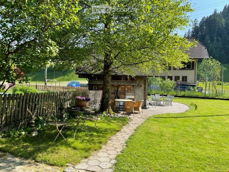 A well-maintained garden area with a wooden fence, a large tree, outdoor seating including tables and chairs, and a paved pathway leading to a house.
