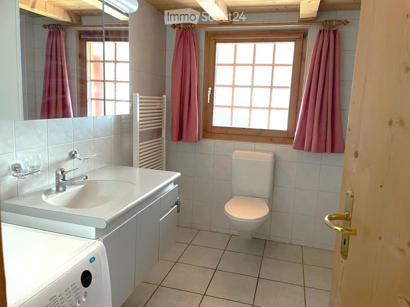 Bathroom with a white sink, toilet, mirror, towel rack, and pink towels. The room has tiled walls and a window with wooden frame and grid pattern.