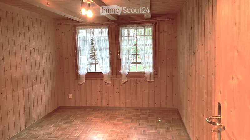Empty room with wooden walls and floor, two windows with white curtains, and a door with a silver handle.