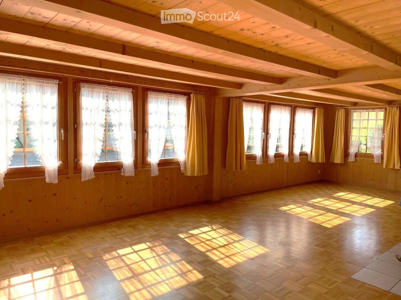 Empty room with wooden walls, wooden ceiling beams, and wooden flooring. Large windows with white and yellow curtains allow natural light to fill the space.