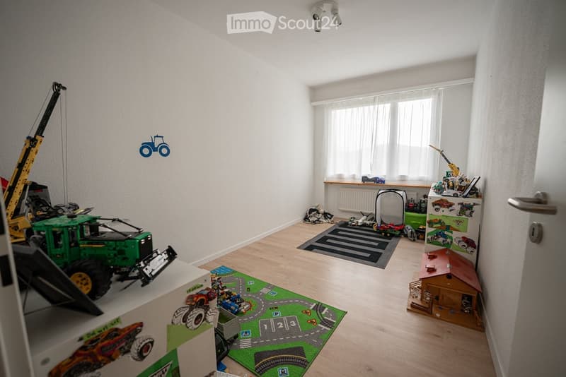 Bright, child-friendly room with wooden flooring, large window with white blinds, and various toy vehicles and play structures.