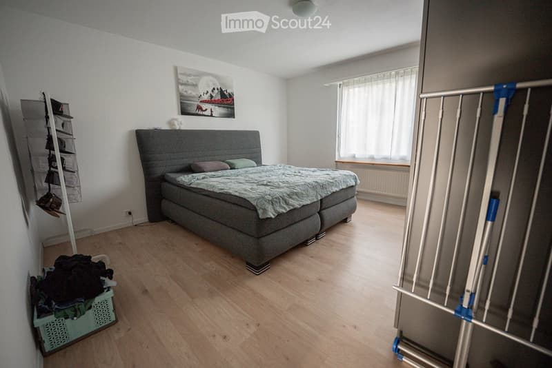 A modern bedroom with a large grey bed, two pillows, a duvet, a nightstand, a picture frame on the wall, a storage unit, and a laundry basket on the floor.