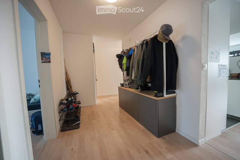 A well-lit hallway with wooden flooring, a coat rack with jackets and hats, a small storage cabinet, and a shoe rack with shoes and bags. A glimpse of a bedroom and a kitchen area is visible.