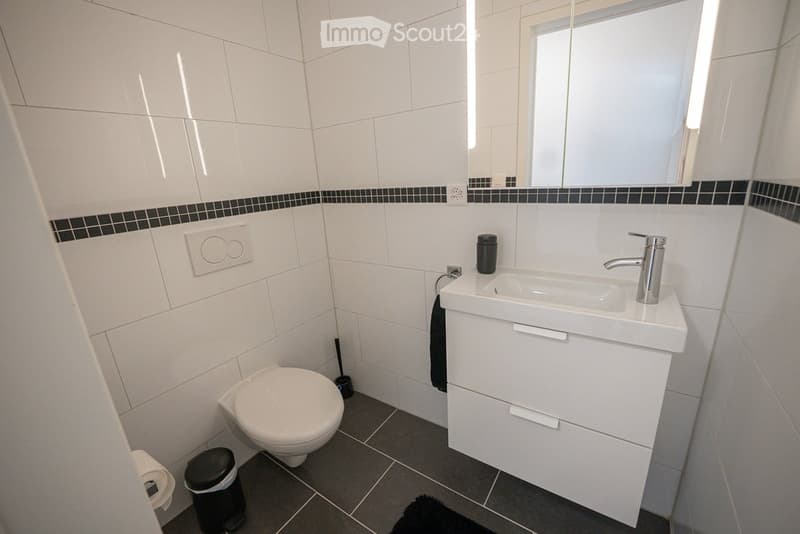 Modern bathroom with white tiles, a toilet, a sink with a chrome faucet, a mirror, and a small black trash bin.