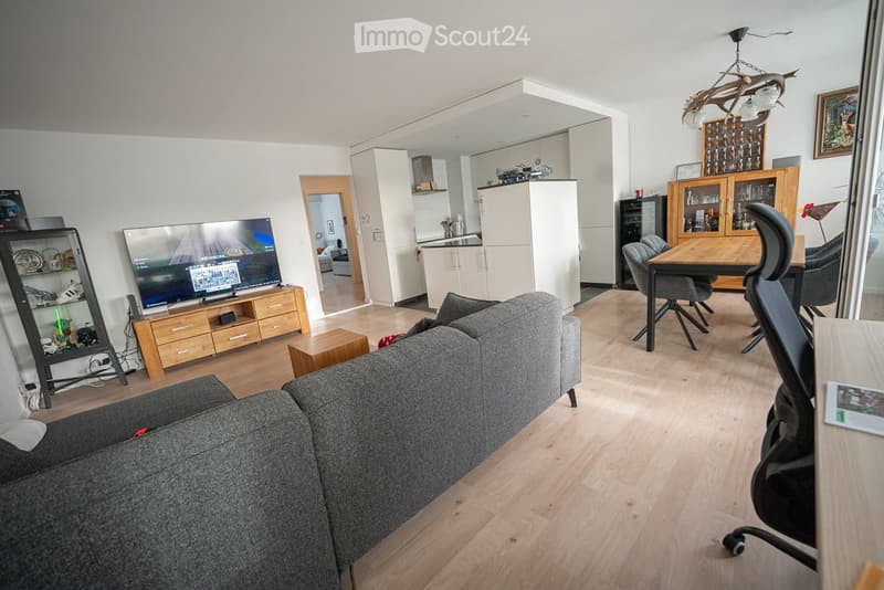 Modern living room with a grey sectional sofa, coffee table, flat-screen TV on a wooden stand, dining area with a wooden table and chairs, and a cabinet with glass doors.