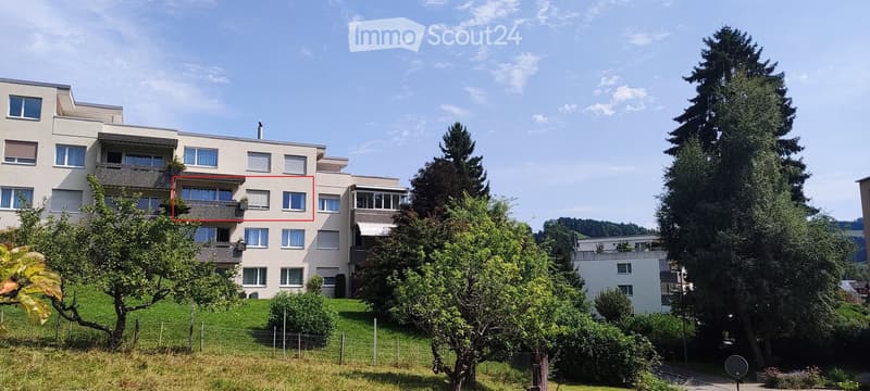 Two-storey apartment building on a raised ground floor with a green hillside in the background.