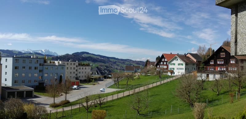 A small hillside village with mountain views, various buildings, a driveway with cars, a slope, and green grass