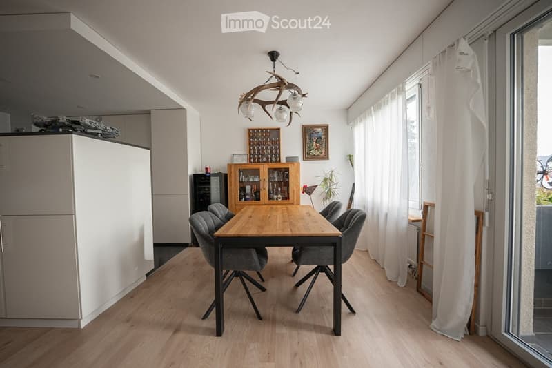 Bright dining area with a wooden dining table and four grey chairs, white cabinets, a refrigerator, and a chandelier with antler design.
