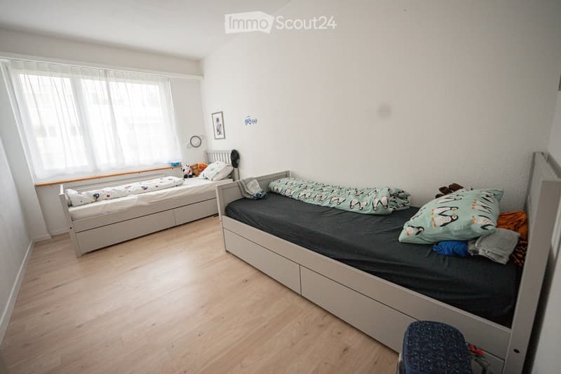 A small, well-lit bedroom with two single beds, one with a penguin-patterned duvet and the other with a colorful blanket. The room features light wooden flooring, white walls, and a large window with sheer curtains.