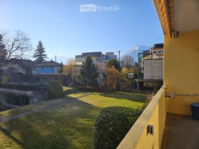 Balcony with a view of a well-maintained garden with green grass, bushes, and trees, surrounded by buildings and mountains in the distance.