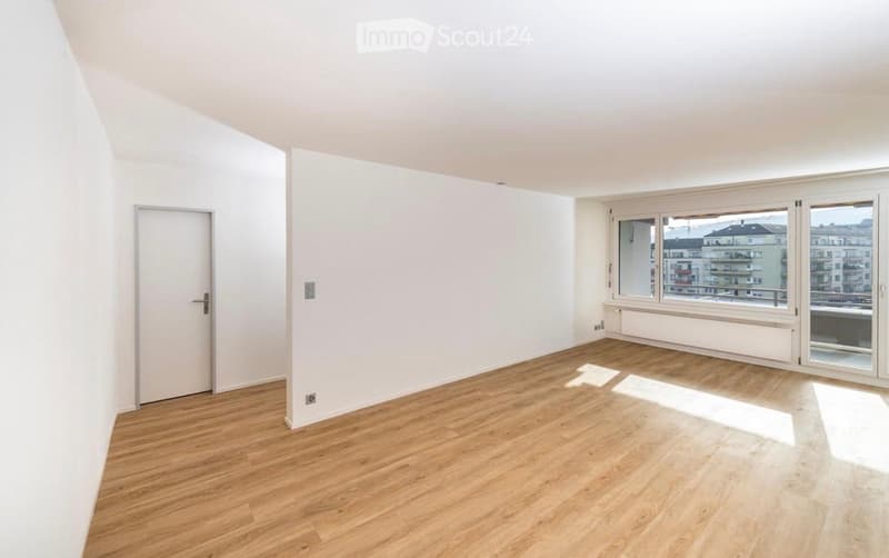 Empty room with light wooden flooring, white walls, and large windows offering a city view. No furniture or equipment present.
