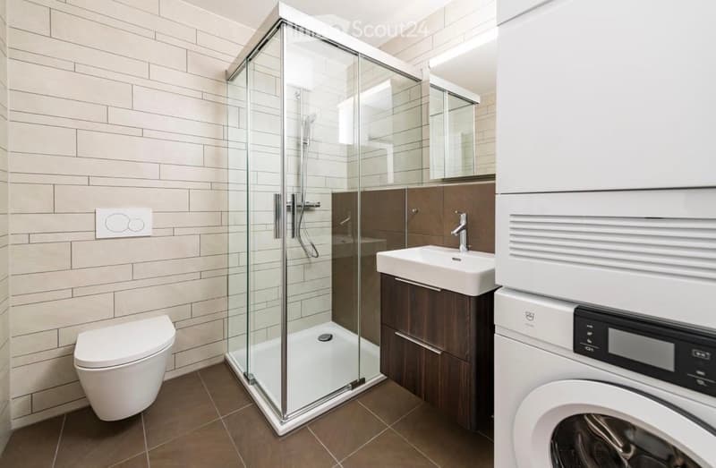 Modern bathroom with a glass-enclosed shower, toilet, sink with wooden cabinet, and a washing machine. The walls are tiled in light beige, and the floor is brown tiled.