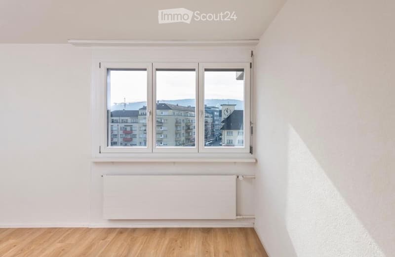 Empty room with light wooden flooring, white walls, and a large window offering a view of the cityscape.