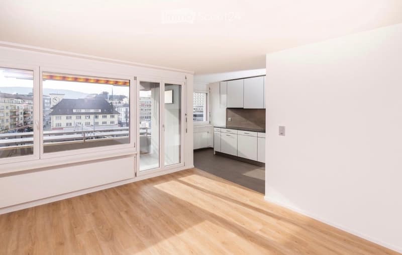 Bright, open living space with wooden flooring, large windows offering a city view, and a modern kitchen area with white cabinets and a countertop.