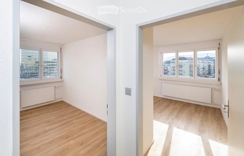 Two adjacent unfurnished rooms with light wooden flooring, white walls, and large windows offering city views. Both rooms feature radiators and electrical outlets.
