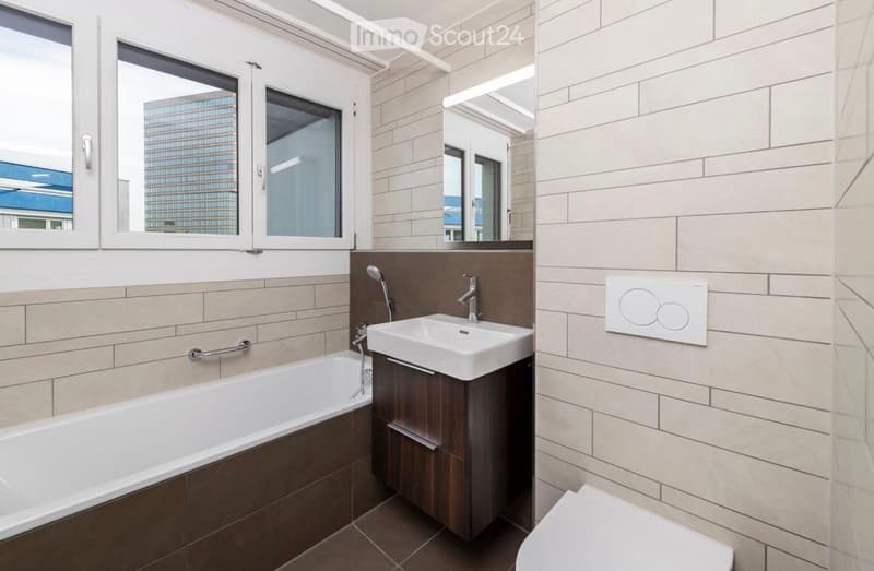 Modern bathroom with a bathtub, sink, and toilet. Features large windows with a city view, beige tiled walls, and a dark wooden vanity.
