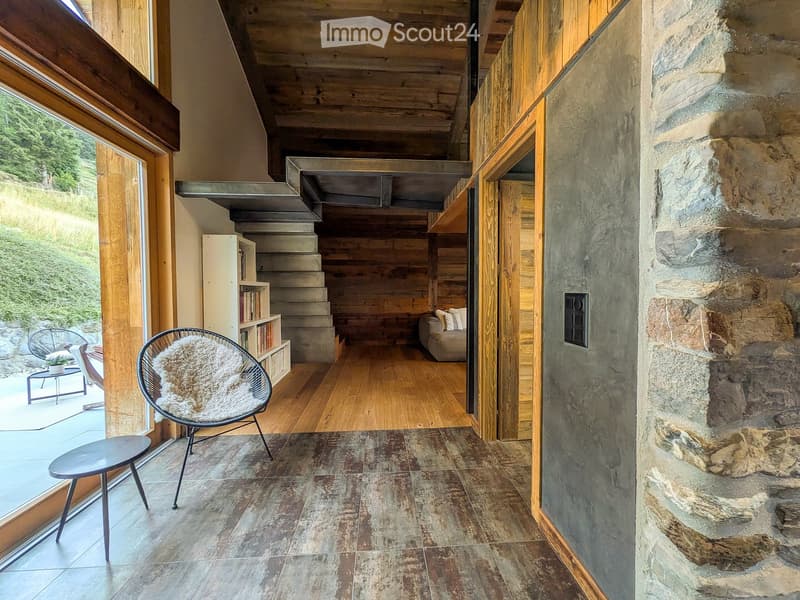 Modern hallway with wooden ceiling, stone walls, and a staircase leading to another level. Features a cozy seating area with a wicker chair, a small table, and a sofa in the background.