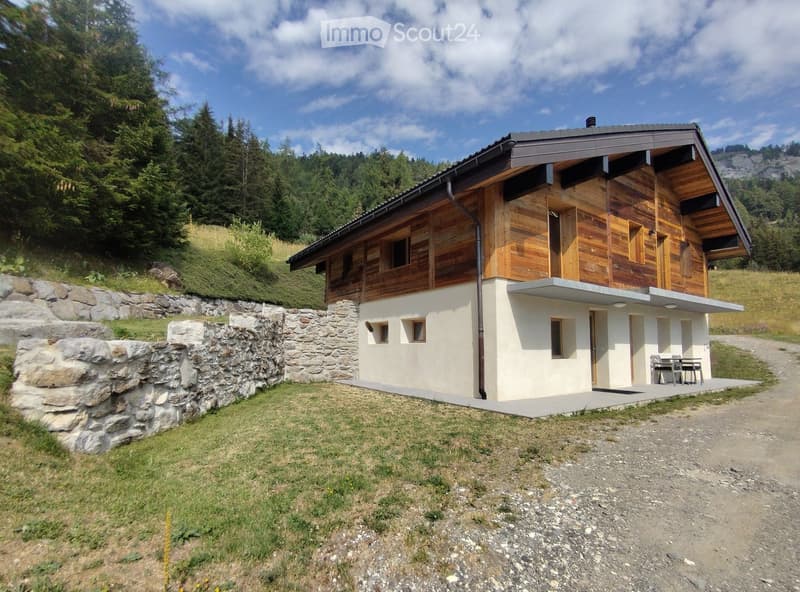 Two-story wooden house with a stone lower level, situated on a raised ground floor with a stone retaining wall, surrounded by greenery and a gravel path.