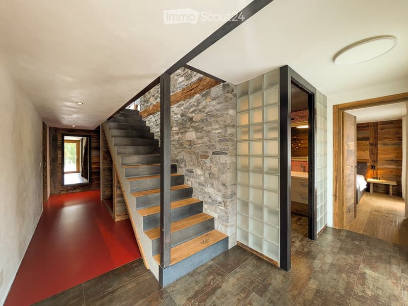 Modern hallway with a stone wall, wooden staircase, glass partition, and red carpeted floor leading to a bedroom area with wooden walls and a bed.