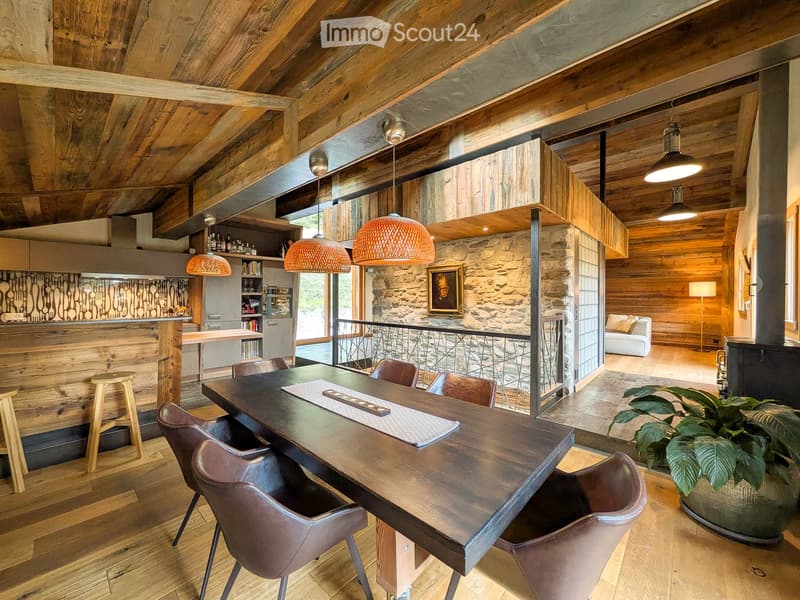Open-plan living and dining area with exposed wooden beams, a large wooden dining table with leather chairs, a bar area with stools, a white sofa, pendant lights, and a stone wall accent.