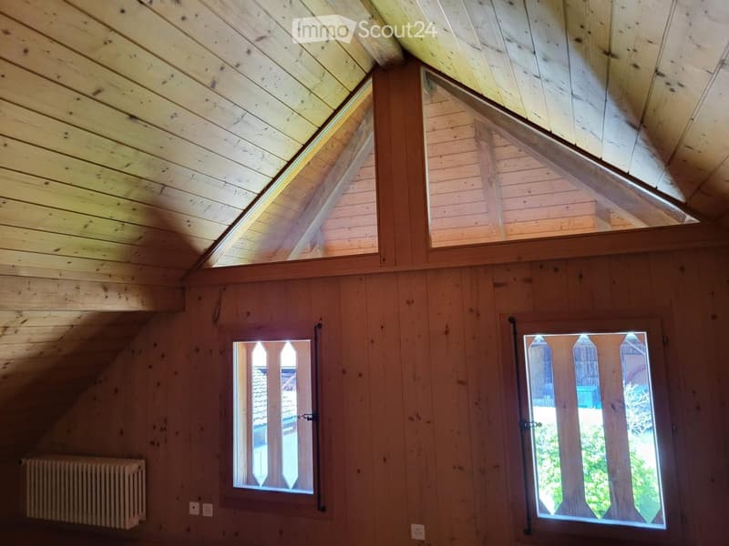 An attic space with wooden walls and ceiling, featuring two windows with wooden frames and a radiator on the left side.
