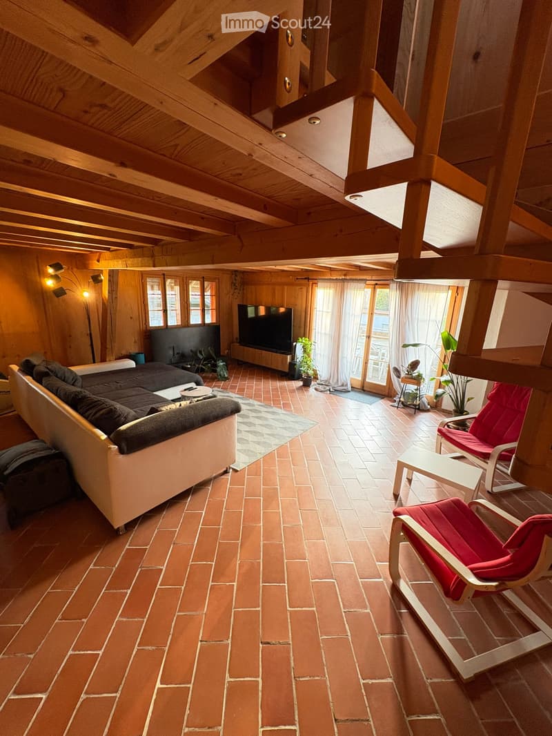 A cozy living room with exposed wooden beams, brick flooring, and large windows. Features a sectional sofa, a red armchair, a coffee table, a television, and a floor lamp.