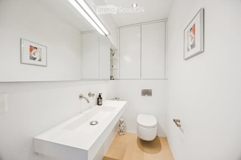 Modern bathroom with white walls, a large sink, a toilet, a mirror, and framed artwork. Features a sleek, minimalist design with a beige tiled floor.