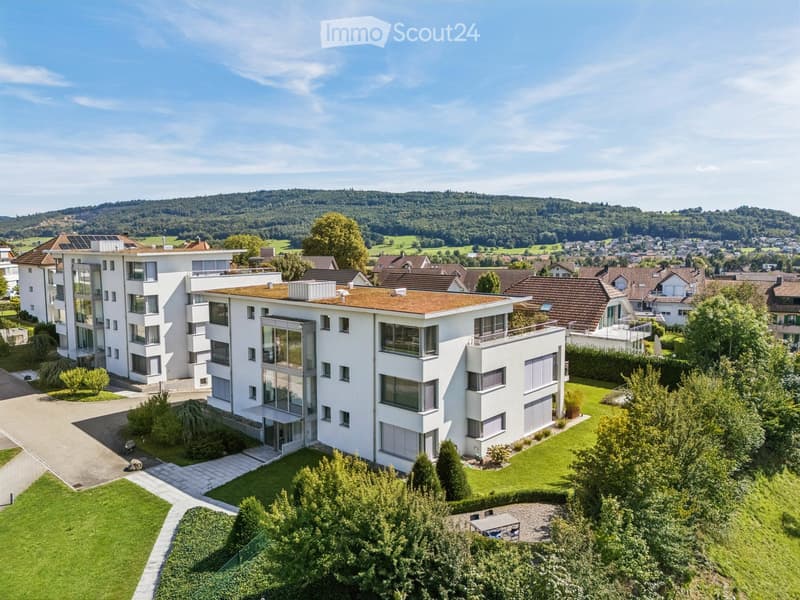 Modern white multi-story building with large windows and a flat roof, surrounded by a well-maintained garden and trees, with a scenic view of a hilly landscape in the background.