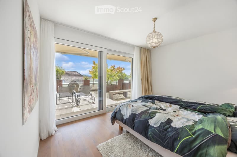 Bright bedroom with wooden flooring, large sliding glass doors leading to a balcony with outdoor seating, a bed with a leafy green duvet, and a hanging light fixture.